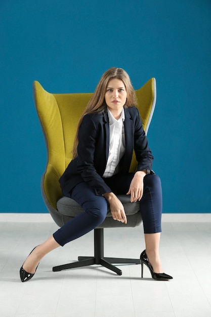 Beautiful fashionable woman in elegant suit sitting in armchair against color surface