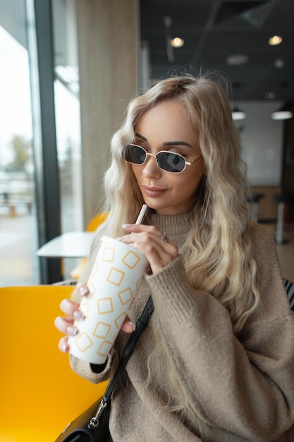 Beautiful fashionable woman with blond curly hair with vintage sunglasses in a beige sweater sits in a cafe holds a paper cup of cola and drinks a drink