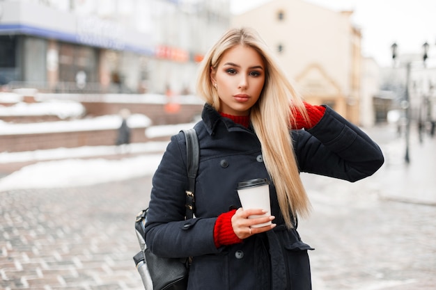 バッグとスタイリッシュなトレンディなコートでコーヒーを飲む美しいファッショナブルな女性が街を旅しています