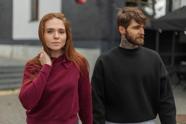 Photo beautiful fashionable young couple a redhaired girl in a hoodie and a handsome bearded guy in fashion outfit walks on the street