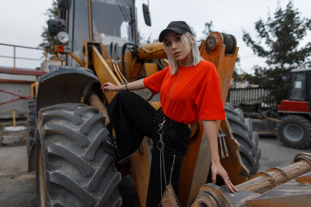 ファッションオレンジTシャツの黒い帽子と建設機械の近くに黒い流行のスニーカーと黒いズボンを持つ美しいファッショナブルな若いスタイリッシュな女性