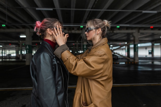 Beautiful fashionable young woman couple in fashionable leather jacket with stylish sunglasses on a parking in the city.  girl puts glasses on her friend outdoors