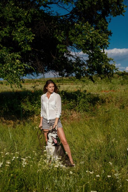 Bella giovane donna alla moda in pantaloncini di jeans e una camicetta bianca in posa nel parco con un husky