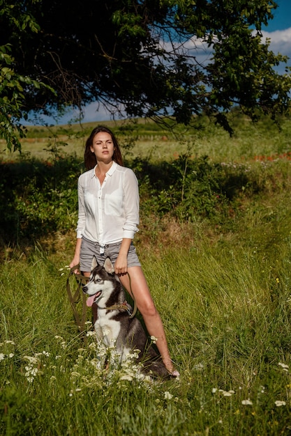 Beautiful fashionable young woman in denim shorts and a white blouse posing in the park with a husky