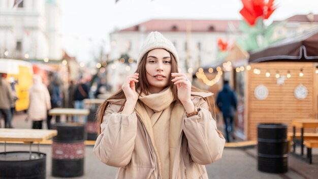 Beautiful fashionable young woman model in winter collection outerwear with a knitted hat in a stylish beige jacket on a city background