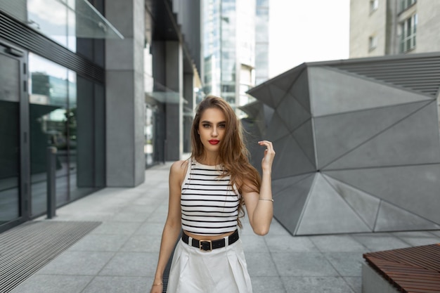 Beautiful fashionable young woman with red lips in stylish casual summer clothes walks in the city Urban female style look