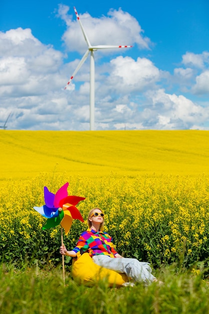 Beautiful female in 90s stylish shirt with pinwheel sitting in rapeseed field