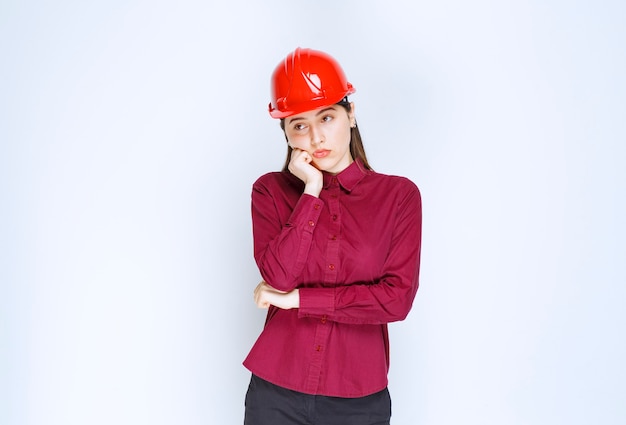 Beautiful female architect in red helmet standing and thinking hard. 