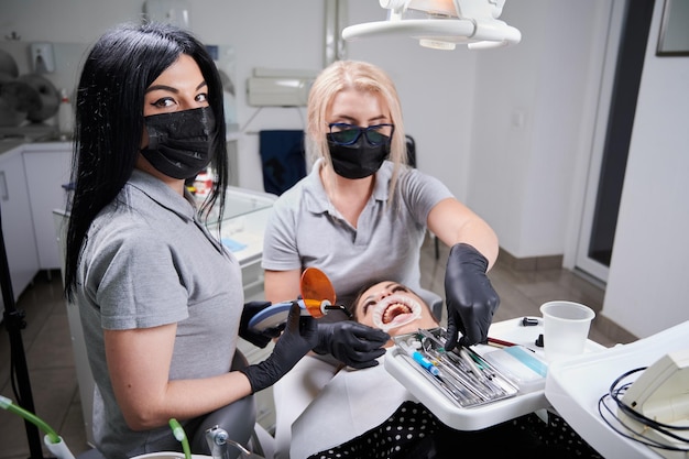 医師が歯科医院で患者の歯にブラケットを取り付けている間、カメラを見ている医療マスクの美しい女性アシスタント 歯科と矯正治療のコンセプト