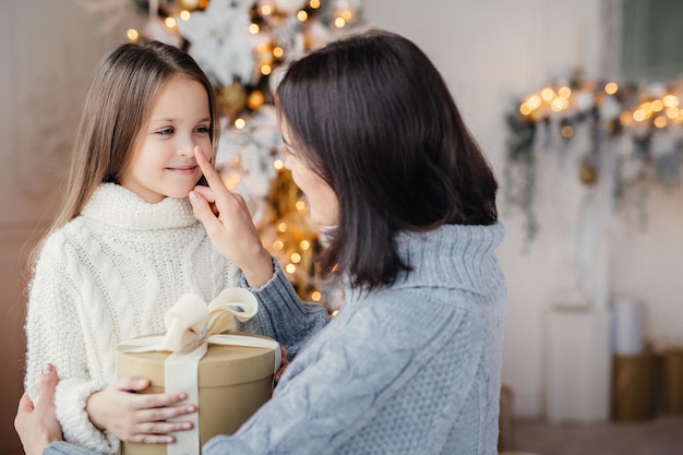 長い髪の美しい女児は白い暖かいセーターを着ており、クリスマスにプレゼントを受け取って喜んで母親の目に見えます家族の輪で冬の休暇を祝います幸せな母と娘