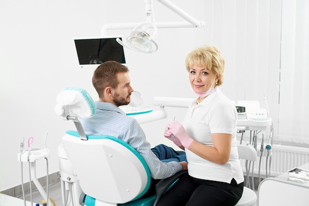 Beautiful female dentist is looking at the camera holding medical instruments and being ready to cure the teeth of young male client of the dentistry.