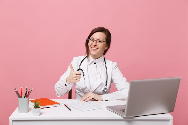 Beautiful female doctor sits at desk works on computer with medical document in hospital isolated on pastel pink wall background. Woman in medical gown glasses stethoscope. Healthcare medicine concept