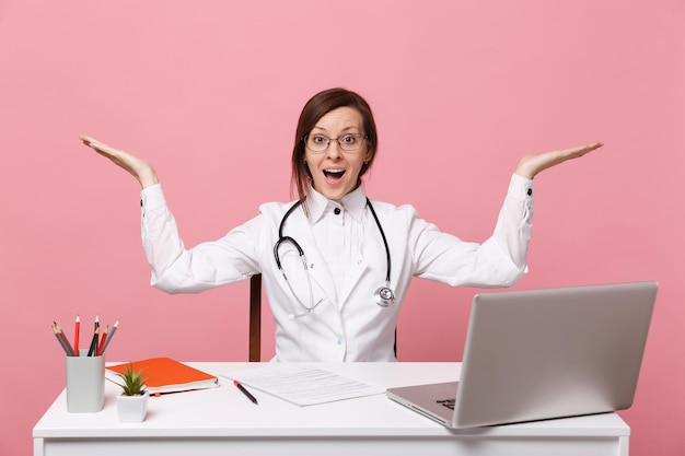 Beautiful female doctor sits at desk works on computer with medical document in hospital isolated on pastel pink wall background. Woman in medical gown glasses stethoscope. Healthcare medicine concept
