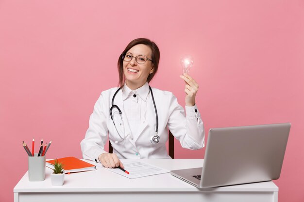 Beautiful female doctor sits at desk works on computer with medical document in hospital isolated on pastel pink wall background. Woman in medical gown glasses stethoscope. Healthcare medicine concept