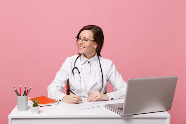 Beautiful female doctor sits at desk works on computer with medical document in hospital isolated on pastel pink wall background. Woman in medical gown glasses stethoscope. Healthcare medicine concept