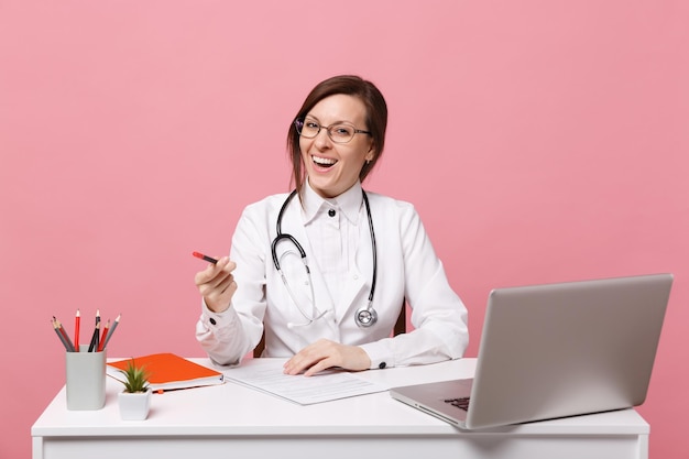 Beautiful female doctor sits at desk works on computer with medical document in hospital isolated on pastel pink wall background. Woman in medical gown glasses stethoscope. Healthcare medicine concept