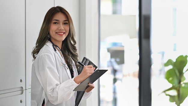 Beautiful female doctor in white coat with stethoscope holding her patient chart and smiling to camera