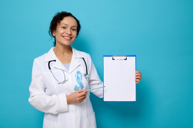 Beautiful female doctor in white medical gown and blue awareness ribbon smiles toothy smile looking at camera and shows a copy space on empty blank paper sheet on clipboard. World Diabetes Day concept