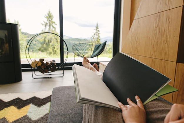 Beautiful female feet and book on background of fireplace and\
big window in modern chalet cozy home