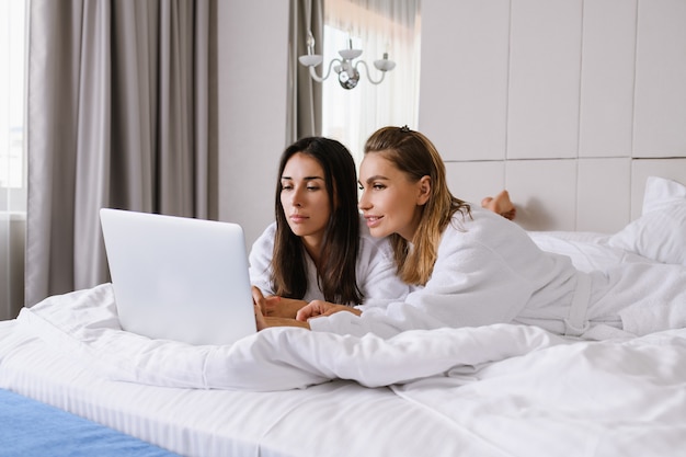 Beautiful female friends at luxury white hotel appartment laying in bed in bathrobes, look at laptop screen, shopping, chatting, leisure, using services online