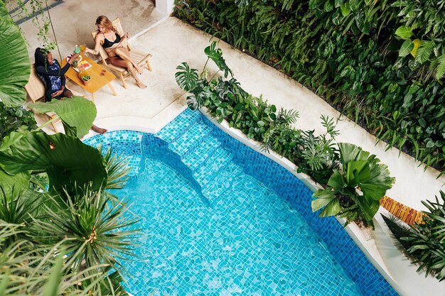 Beautiful female friends sitting at table by swimming pool of spa resort, eaitng fresh fruits and drinking cocktails, view from above