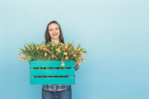 Beautiful female gardener holding box with tulips on blue background