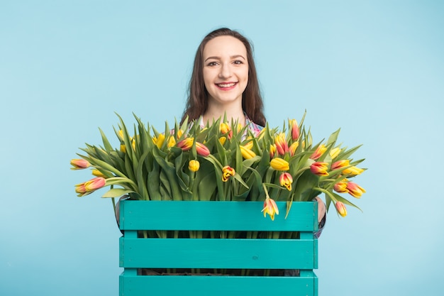 Beautiful female gardener holding box with tulips on blue surface