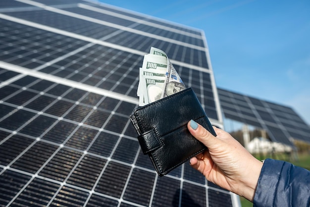 美しい女性の手は、彼女がソーラー パネルのグリーン電力ビジネス購入の太陽の概念の近くに保持しているドル札で黒い財布を保持しています。