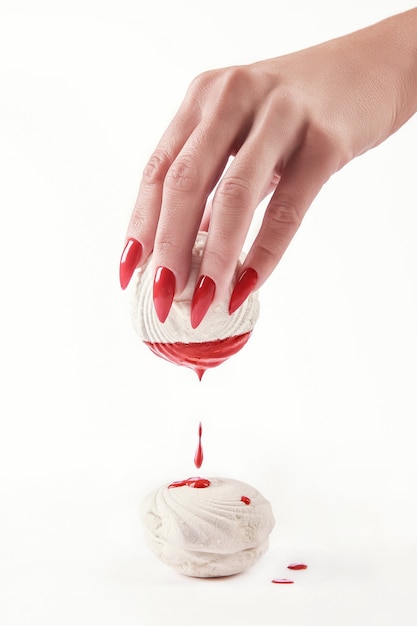 Beautiful female hand with red manicure holding a marshmallow, with a dripping red paint on the marshmallows. White background. Nail extension. Manicure, Spa salon. Creative, advertising. Relax.