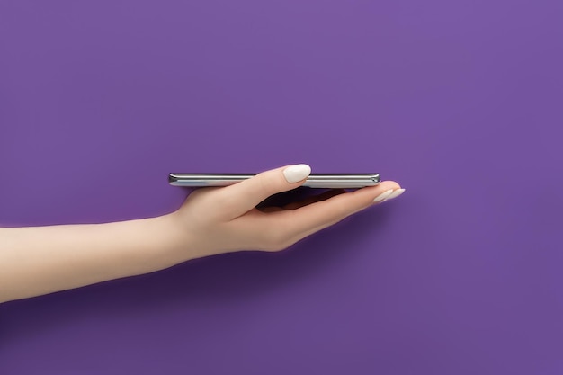 Beautiful female hand with white manicure holding smartphone of silver color on purple background