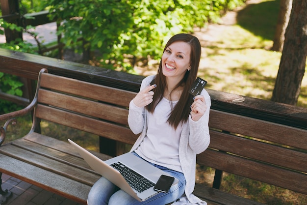 Beautiful female holding credit card with copy space. Woman sitting on bench working on modern laptop pc computer, mobile phone in street outdoors on nature. Mobile Office. Freelance business concept.