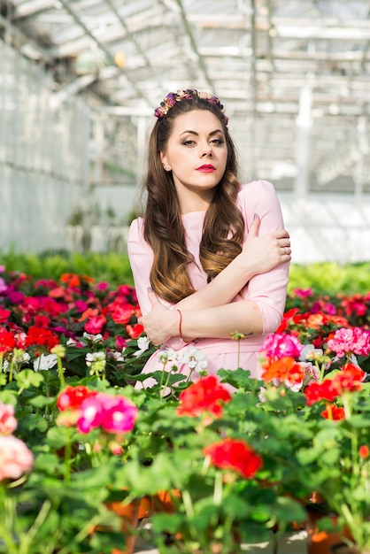 Beautiful female model dressed in a long pink dress poses among the many flowers in the greenhouse