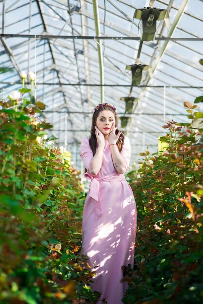 Красивая женская модель, одетая в длинное розовое платье, позирует среди множества цветов в оранжерее