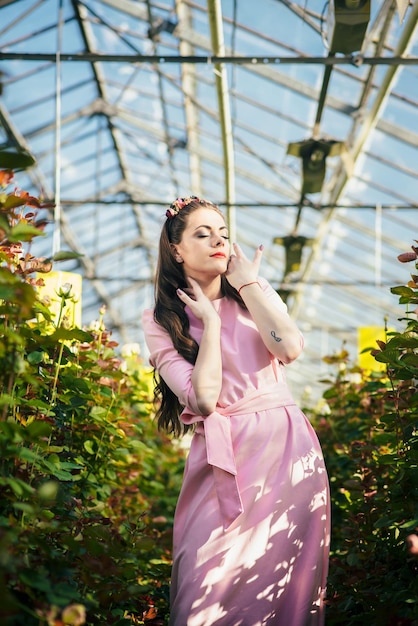 Beautiful female model dressed in a long pink dress poses among the many flowers in the greenhouse