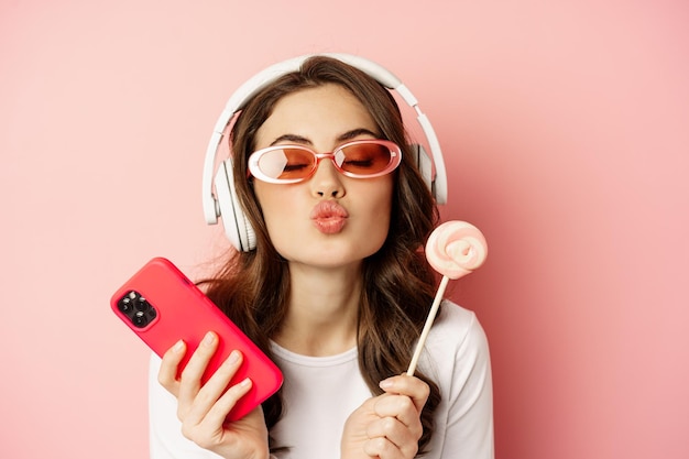 Красивая женская модель слушает музыку в наушниках, держит леденец и мобильный телефон, позирует в солнцезащитных очках, стоит на розовом фоне