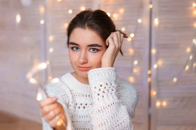 Beautiful female portrait of a young girl in a white sweater on a background of garlands and bokeh. Cozy festive atmosphere