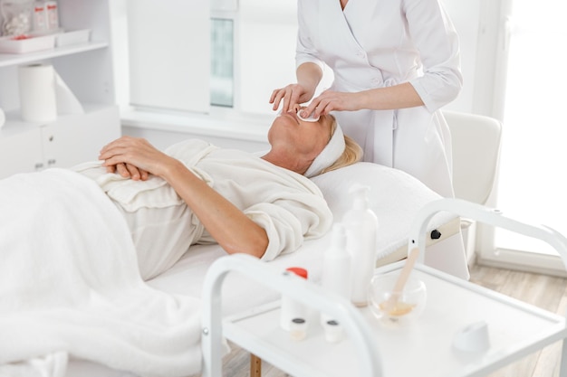 Beautiful female professional doctor is cleaning a skin of mature blond woman in modern cosmetology center