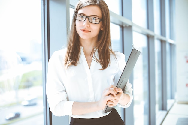 Beautiful female specialist with clipboard standing in modern office and smiling charmingly Working on design data analysis plan strategy Business people concept