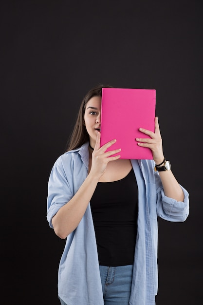 Photo beautiful female student in denim shirt with long hair, on black wall, peeking out from behind book