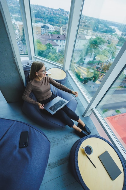 A beautiful female student works on a laptop while sitting on a
bean bag chair in a modern creative office against the background
of panoramic windows break time concept