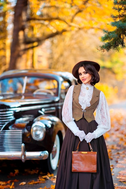 Beautiful female in vintage  dress, lace blouse and hat with veil standing near retro brown car