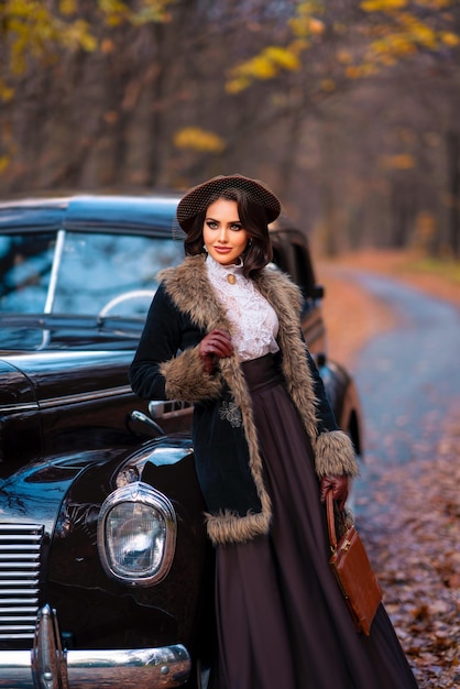 Beautiful female in vintage  dress, lace blouse and hat with veil standing near retro brown car