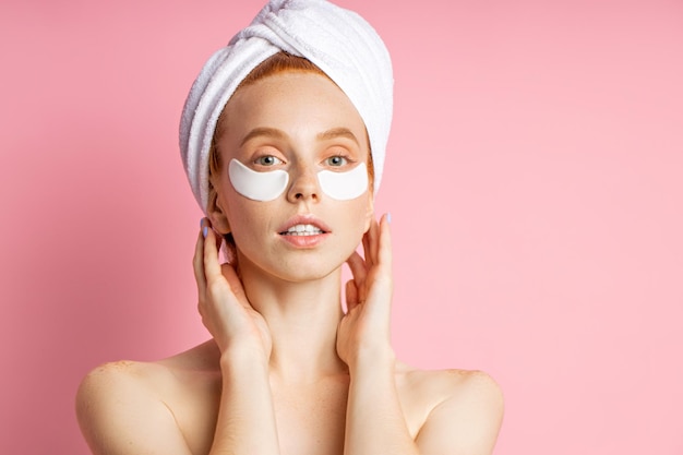 Beautiful female with anti aging, rejuvenating eye patches on face. Image of young gentle redhead woman standing wearing towel on head, taking care of her skin on pink background.