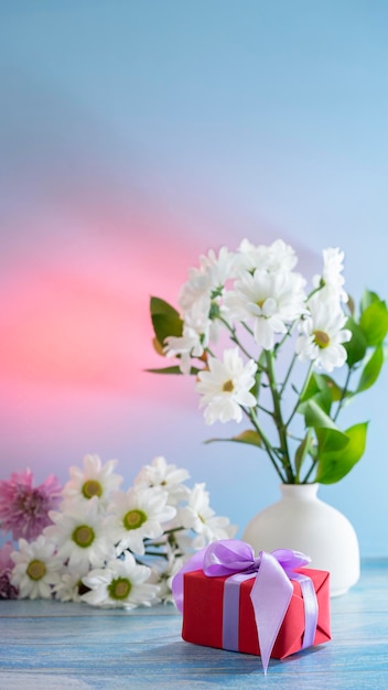 Foto bella natura morta festiva con un regalo in carta rossa, fiori bianchi in un vaso bianco su sfondo blu.