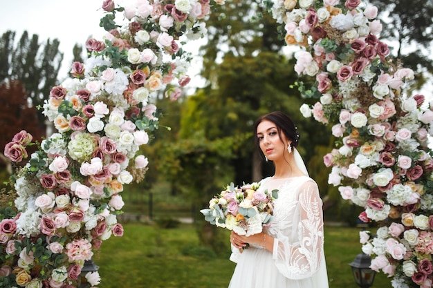 Beautiful fiancee in white wedding dress with bouquet stand near the wedding arch of flowers