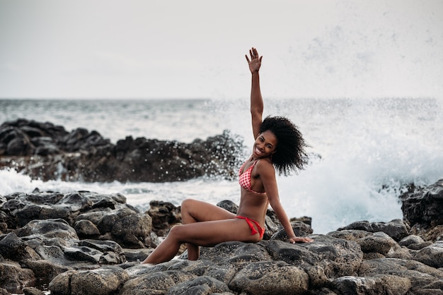 Beautiful fit black woman sitting in rocks in bikini by the seashore