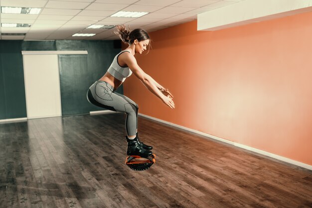 Castana caucasica di bella misura in abiti sportivi che saltano sulle calzature di salti di kangoo in palestra.