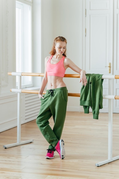 Beautiful fit ginger female dancer has rest near barre in ballet dancing studio focused down with thoughtful expression wears top green trousers and sneakers prepares for performance on stage