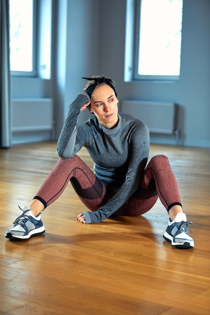 Beautiful fit woman in sportswear posing while sitting on the floor in front of window at gym