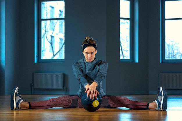 Beautiful fit woman in sportswear posing while sitting on the floor with basketball in front of window at gym. Urban style, healthy girl lifestyle and sport concept. Hipster city life.